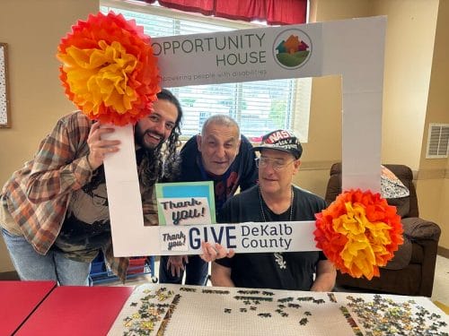 Opportunity House clients and a volunteer working on a puzzle while the volunteer holds a fundraising poster frame.