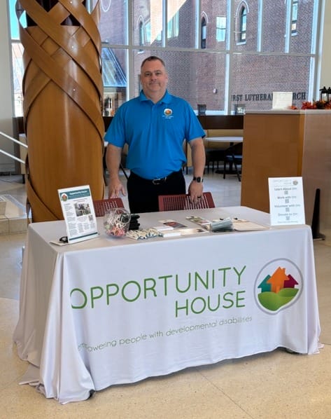 Opportunity House employee in blue shirt standing in front of Opportunity House table.